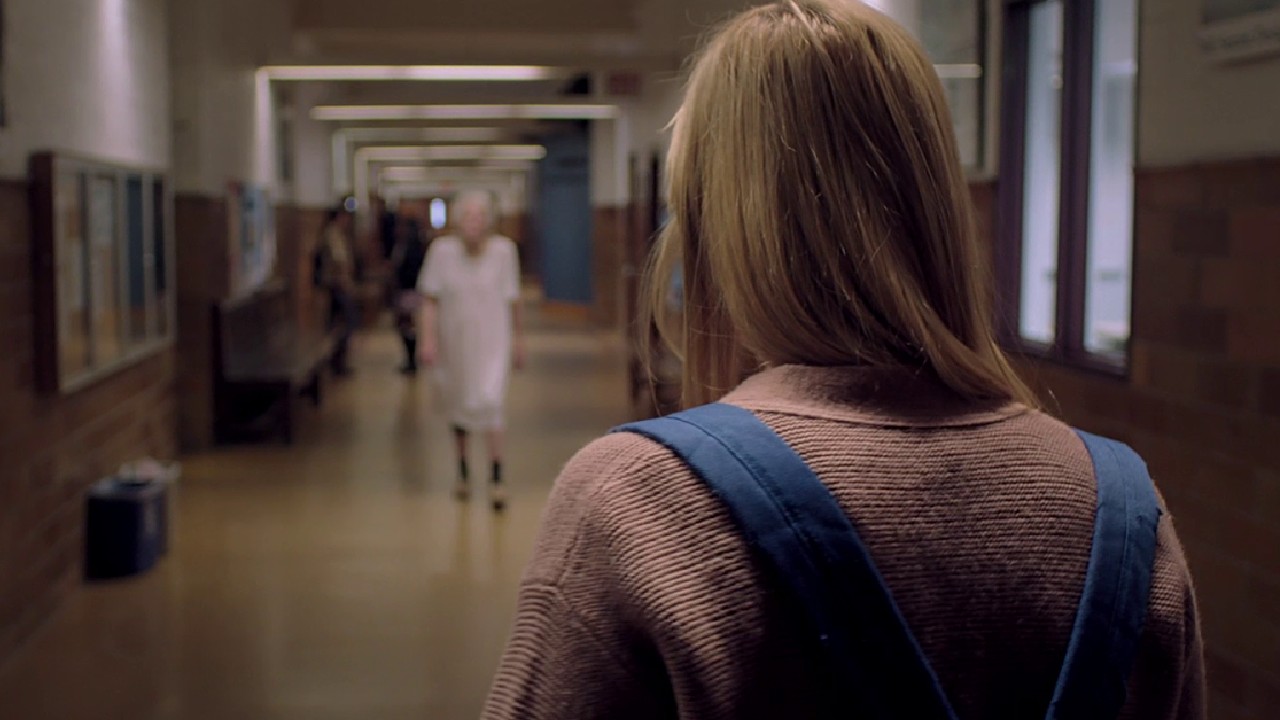 A young woman wearing a backpack stands in a school hallway, facing an elderly woman in a hospital gown walking wo