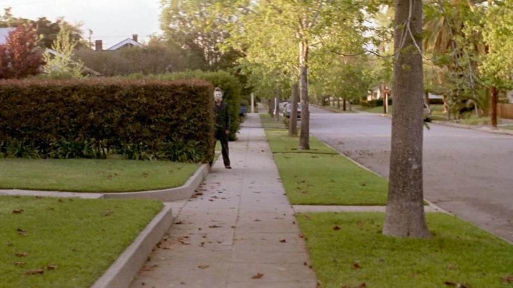 A sunny suburban street with a man in a mask staring from the background