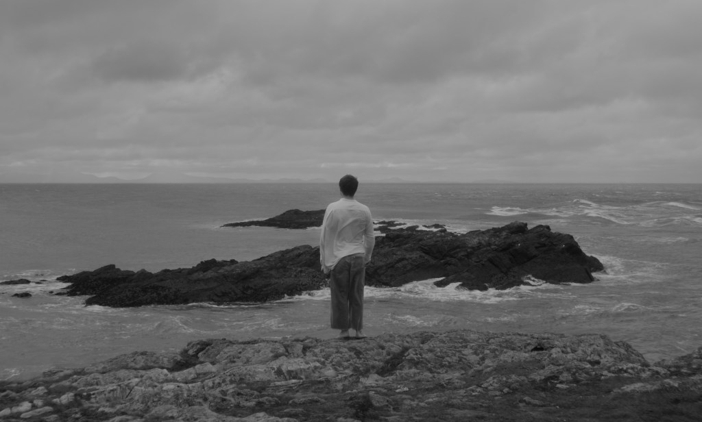 A man with his back to the camera, at the edge of a seaside cliff.