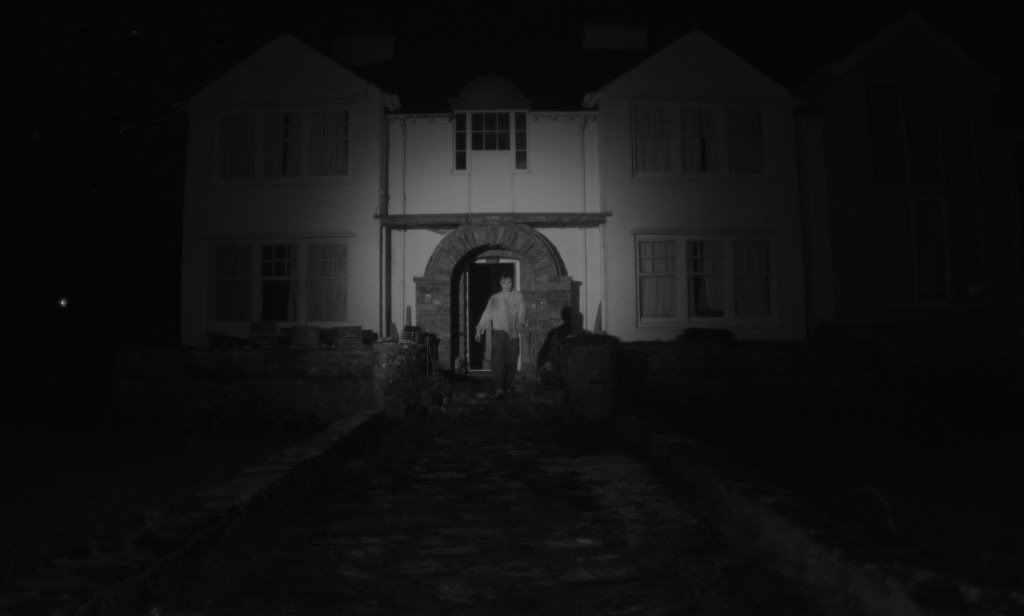 A man stands before a door, surrounded by darkness.